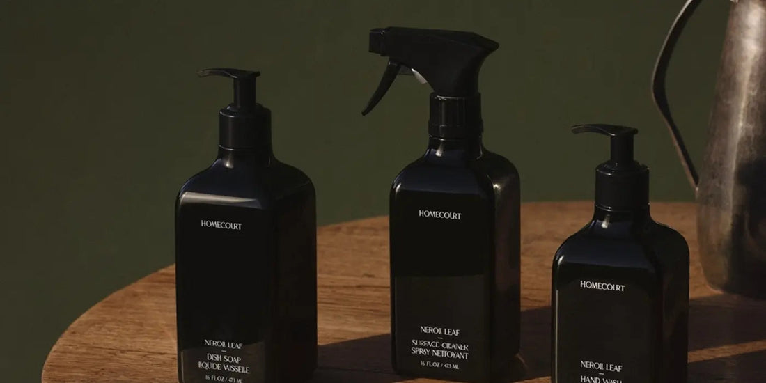 Three-quarter view of three black cleaning bottles with pump and spray tops on a wooden table and dark green background