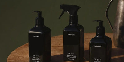 Three-quarter view of three black cleaning bottles with pump and spray tops on a wooden table and dark green background