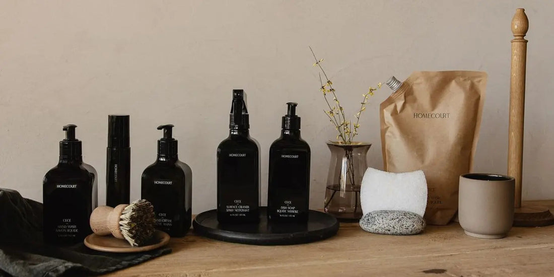 Homecourt cleaning bottles and refill pouch arranged on a wooden table with brushes, sponge, and vase against a neutral wall