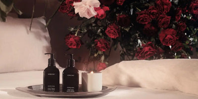 Two dark pump bottles labeled HOMECOURT on a tray with a lit white candle on a bed with pale linens and red roses behind