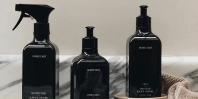 Front view of three Homecourt cleaning bottles with a trigger spray, pump dispenser, and tall soap bottle on a marble countertop
