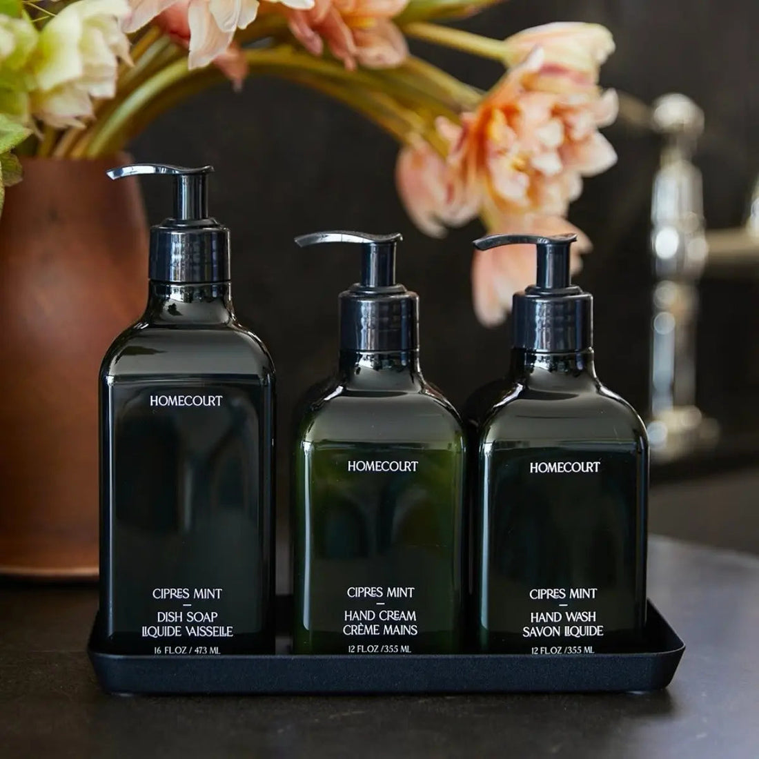 Three Homecourt Cipres Mint pump bottles (dish soap 16oz, hand cream 12oz, hand wash 12oz) on a black tray with pink flowers