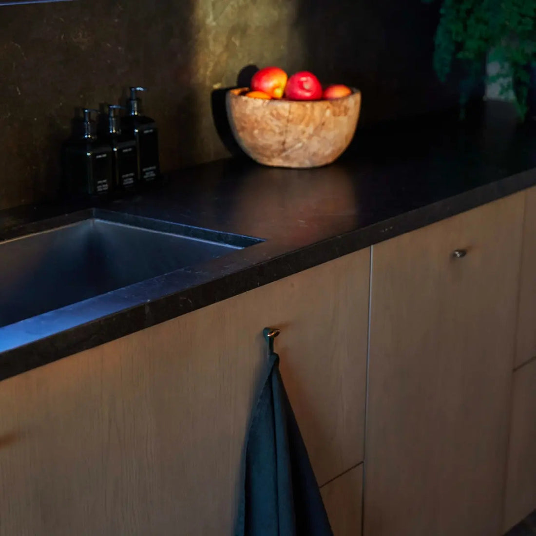 Moody kitchen countertop with dark undermount sink, three black refillable soap dispensers, a wooden bowl of apples, and a hanging towel