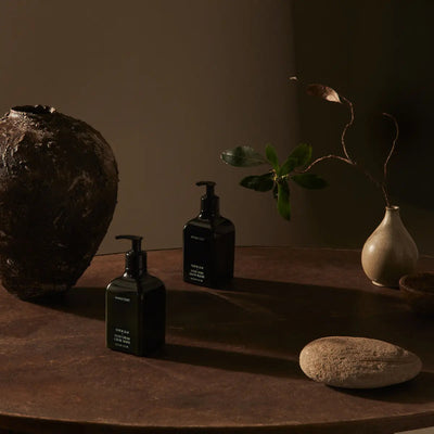 Two dark pump bottles of non toxic body butter on a brown tabletop with clay vases and a green sprig