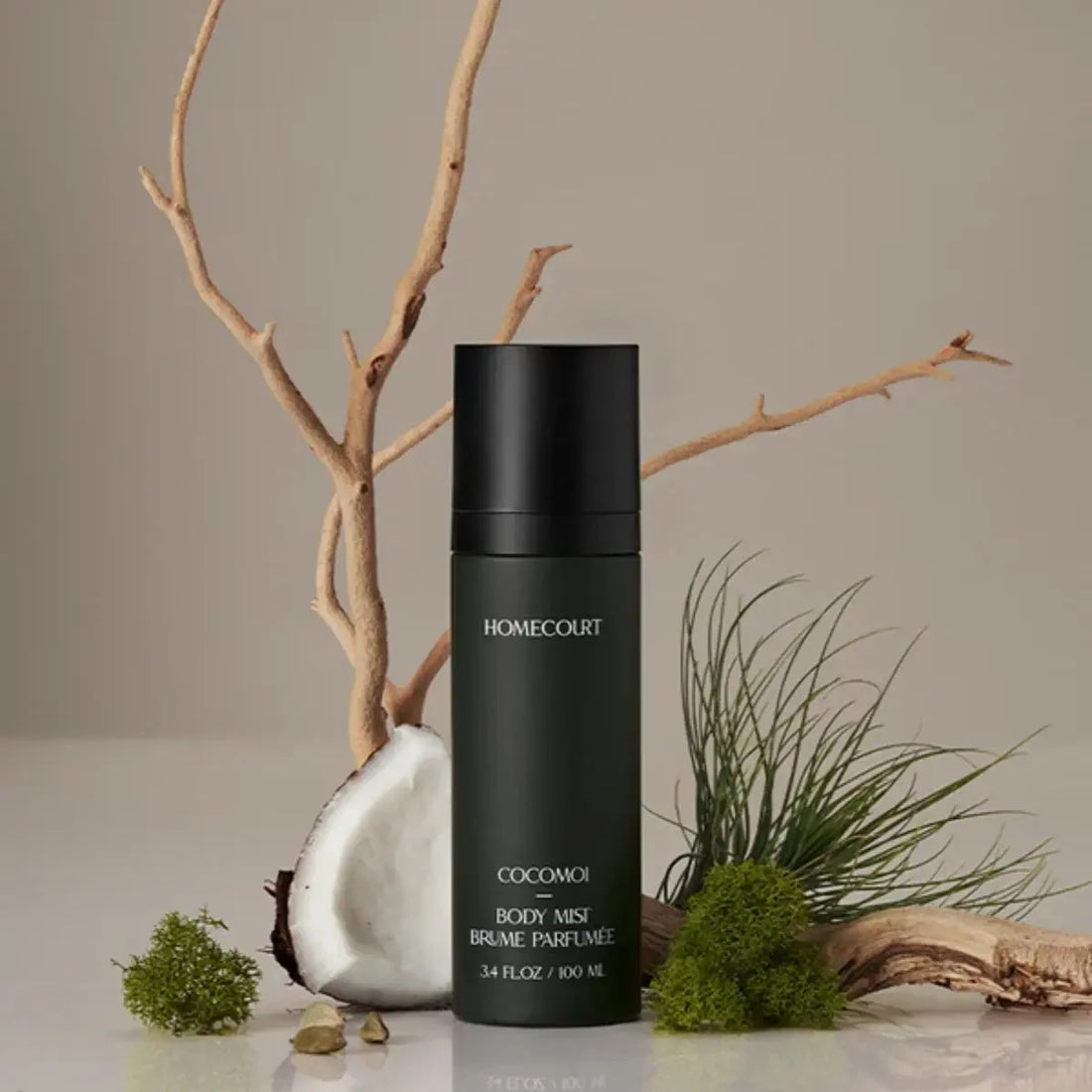 Front view of Homecourt Cocomoi body mist, matte dark green 3.4 fl oz bottle with black cap, styled with coconut and greenery on beige background
