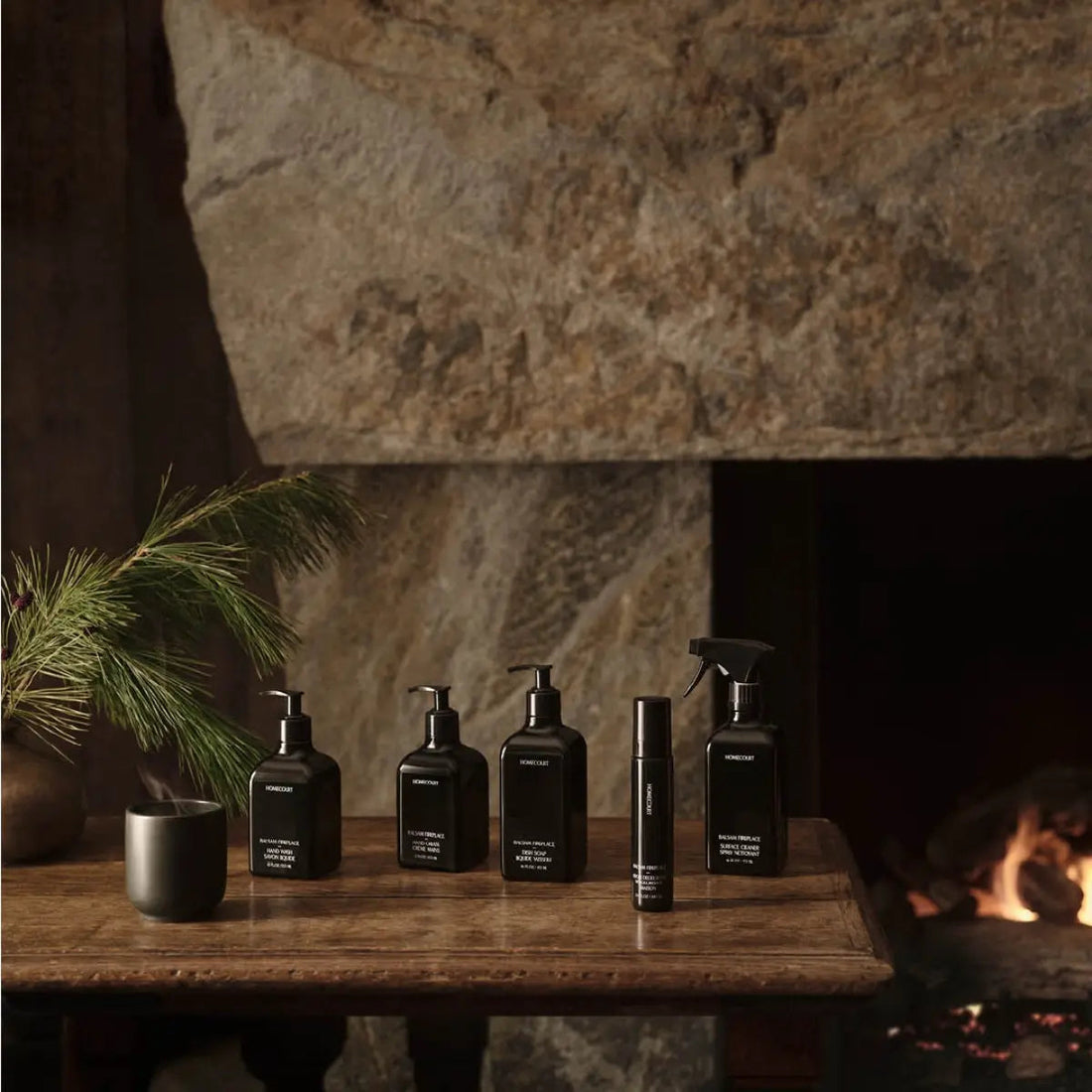 Row of black Homecourt cleaning bottles arranged on a wooden table in front of a stone fireplace with a pine sprig and lit fire