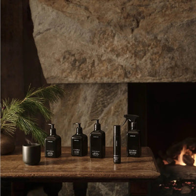 Row of black Homecourt cleaning bottles arranged on a wooden table in front of a stone fireplace with a pine sprig and lit fire