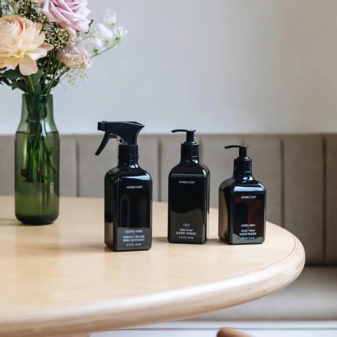 Front view of three black Homecourt bottles with spray and pump tops and minimalist labels on a light wood table with a vase of pink flowers in the background
