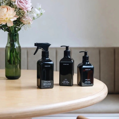 Front view of three black Homecourt bottles with spray and pump tops and minimalist labels on a light wood table with a vase of pink flowers in the background