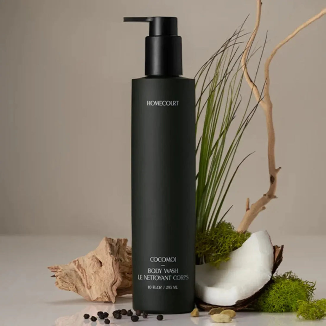 Front view of Cocomoi body wash pump bottle showcasing matte black finish and label with coconut and plant props on beige background