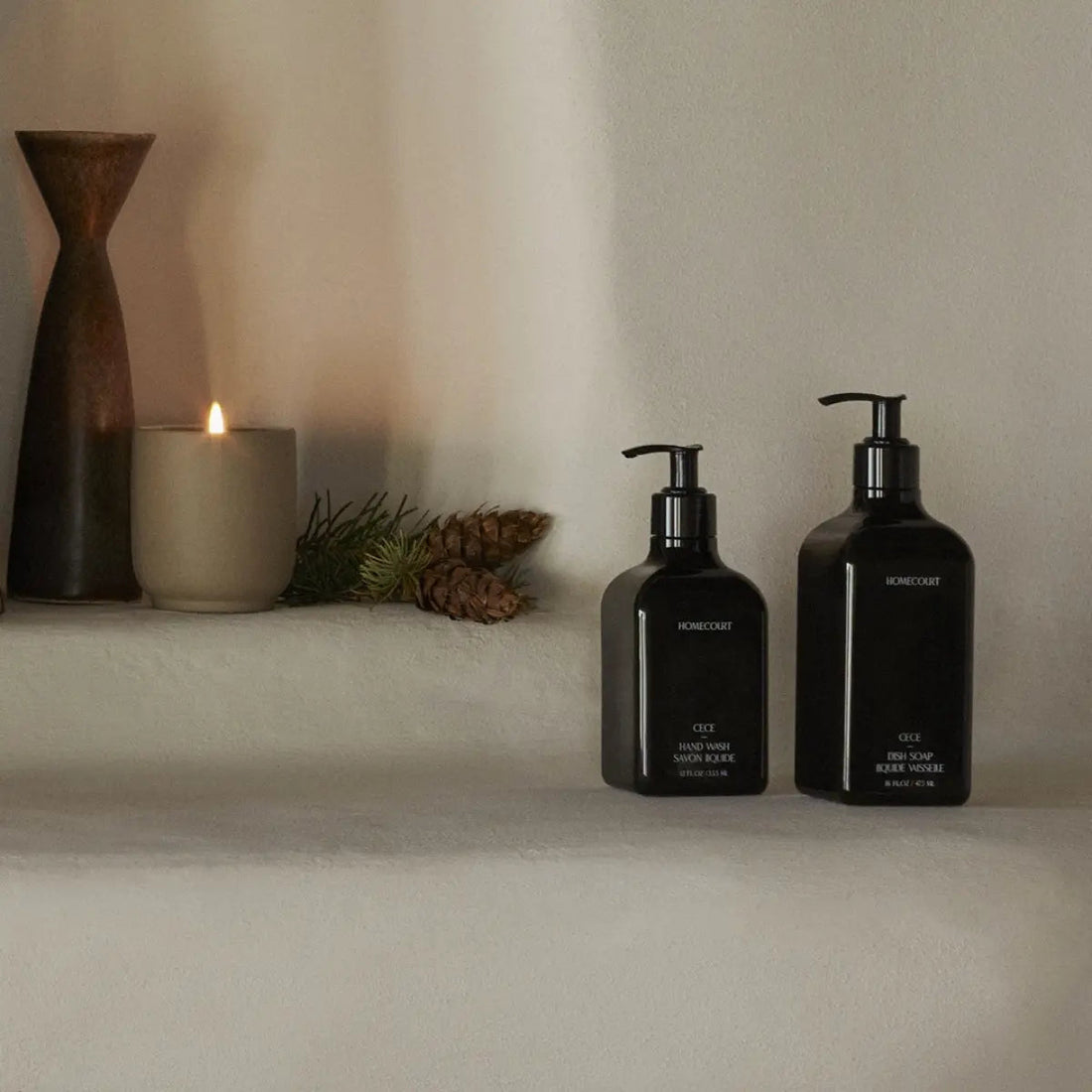 Front view of two Homecourt black pump soap bottles showcasing minimalist labels on a neutral shelf with a lit candle and pinecones in the background