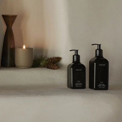 Front view of two Homecourt black pump soap bottles showcasing minimalist labels on a neutral shelf with a lit candle and pinecones in the background