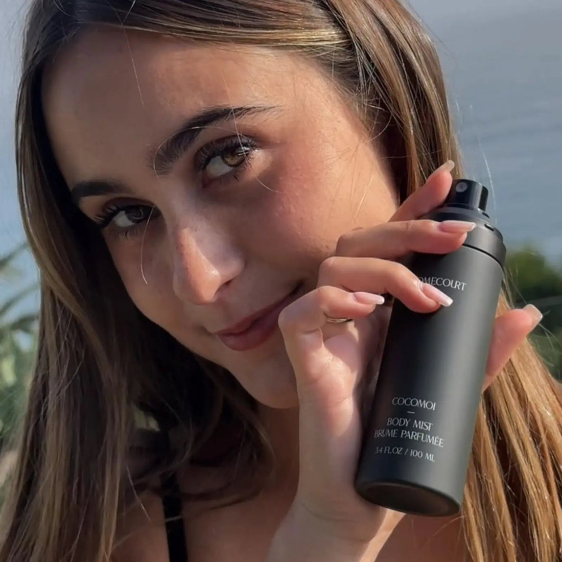 Young woman holding a matte black body mist can near her face outdoors in sunlight