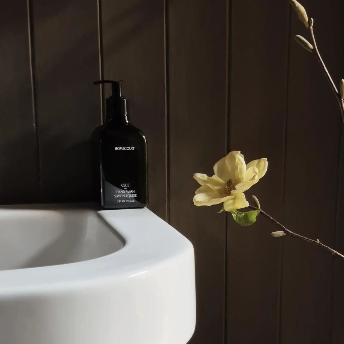 Front view of Homecourt CECE hand wash bottle with pump on white sink, yellow magnolia branch and dark wood paneling