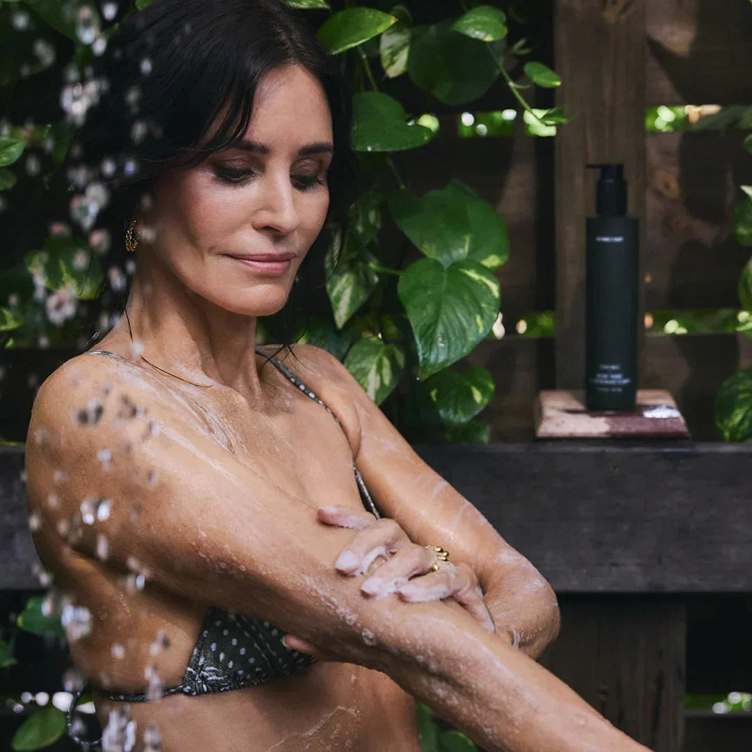 Woman washing her arm with body wash in an outdoor shower, dark pump bottle on a wooden shelf and green leaves in the background