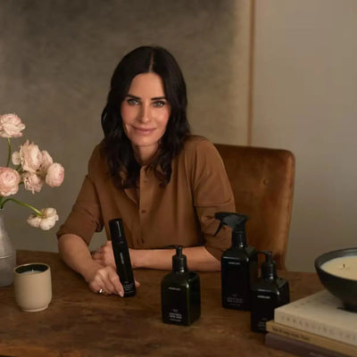 Woman seated at a table with Homecourt black bottles, pale flowers, a candle, and stacked books in a warm room