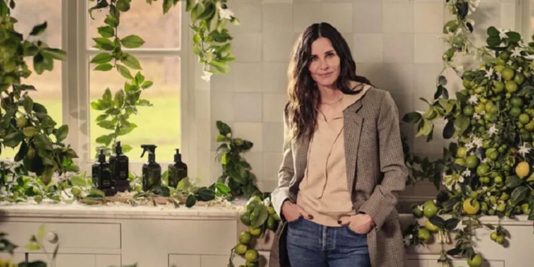 Woman leaning on a kitchen counter beside reed diffuser bottles on a windowsill with green foliage and lemons