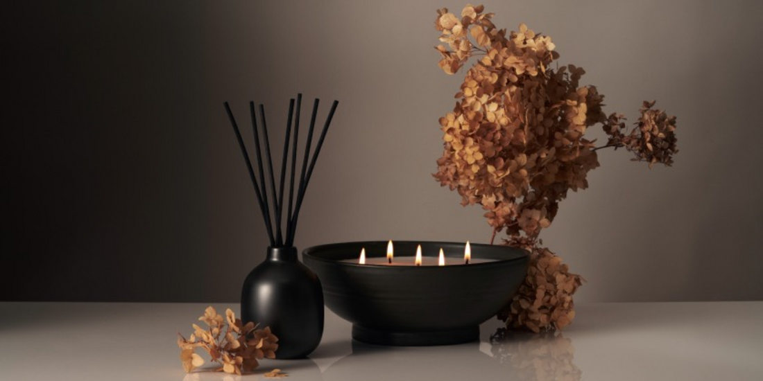Front view of black reed diffuser showcasing tall black reeds next to a low black bowl candle with five lit wicks and dried hydrangea on a neutral background