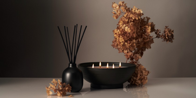 Front view of black reed diffuser showcasing tall black reeds next to a low black bowl candle with five lit wicks and dried hydrangea on a neutral background