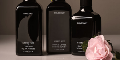 Front view of Steeped Rose dish soap bottle in dark glass with Homecourt label, two matching bottles and a pink rose