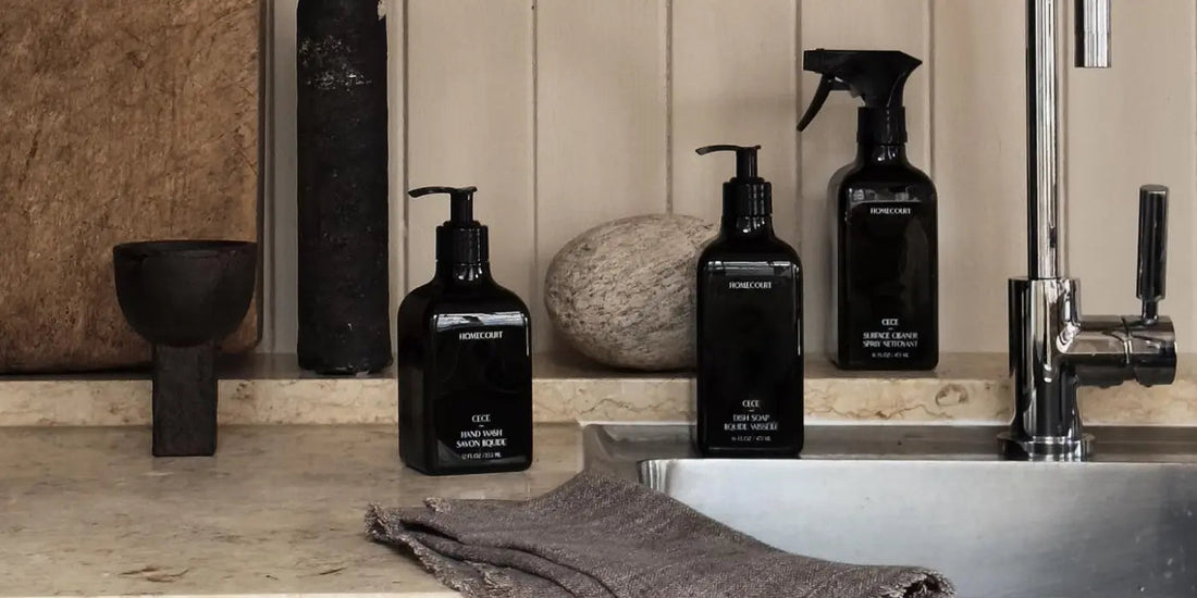 Three black Homecourt cleaning bottles on a marble kitchen counter beside a stainless steel sink with a folded taupe towel