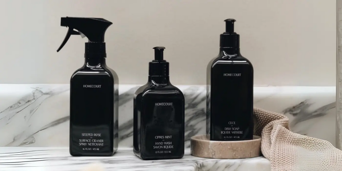 Front view of three Homecourt cleaning bottles, spray, hand wash, dish soap, with black pumps on marble and a beige cloth