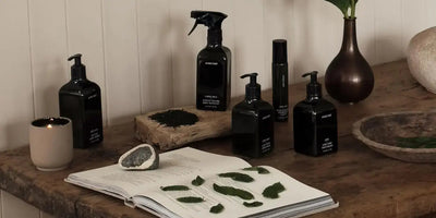 Black glass skincare bottles and a lit candle arranged on a rustic wooden table with an open book and pressed green leaves