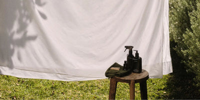 Laundry concentrate bottles and a folded cloth resting on a wooden stool beside a sunlit white sheet hung to dry outdoors