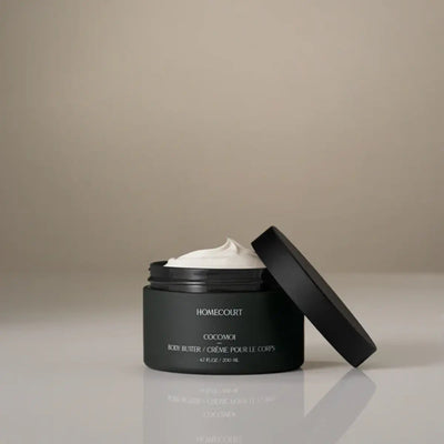 Front view of Homecourt Cocomoi body butter jar with open lid showing whipped cream texture on a neutral beige background