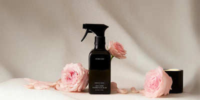 Front view of linen spray bottle with black trigger, surrounded by pink roses and a lit candle on a soft beige background