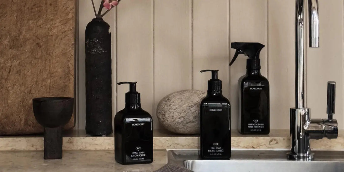 Three black Homecourt cleaning bottles sitting on a marble sink counter beside a chrome faucet and stone decor