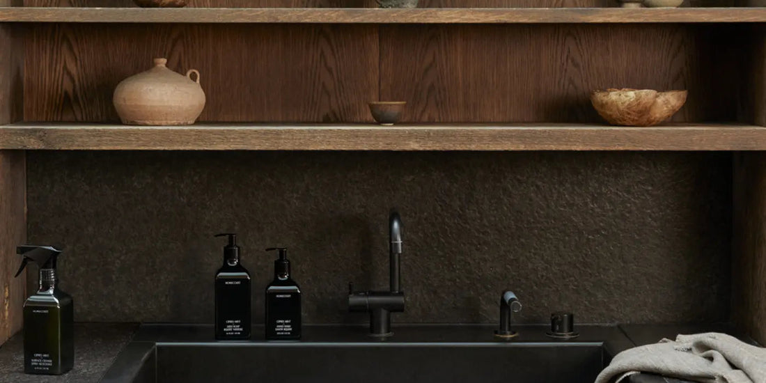 Three dark bottles of plant-derived degreasers sitting on a black kitchen sink with wooden shelves and decorative bowls in the background