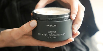 Close-up of Homecourt Cocomoi body butter jar (200 ml) showing creamy texture as hands hold it against a blurred dark background