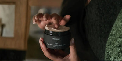 Close-up of Homecourt Cece body butter jar 200 ml held in hands, opened to show creamy texture on fingertip against blurred indoor background