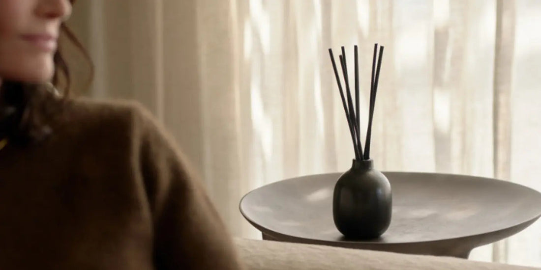 Reed diffuser in a matte black vase sitting on a shallow round tray beside a woman in a brown sweater and sunlit curtains