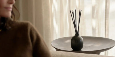 Reed diffuser in a matte black vase sitting on a shallow round tray beside a woman in a brown sweater and sunlit curtains