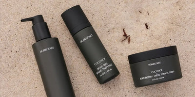 Luxury body care set including a body wash, body mist, and body butter laid flat on a sandy textured surface.