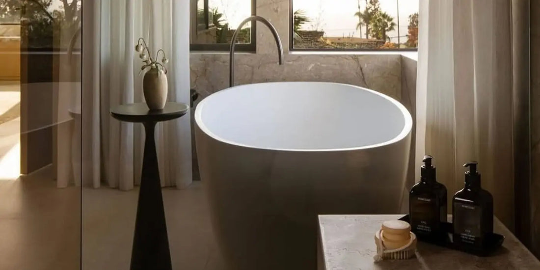 Luxury body wash and body butter bottles displayed on a stone ledge beside a freestanding oval bathtub with a garden view.