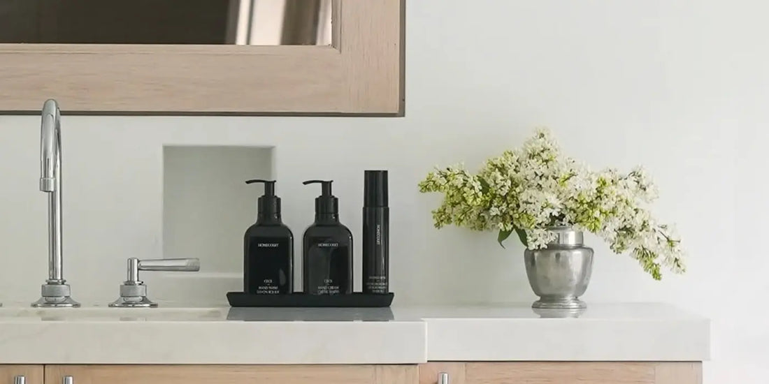 Luxury fragrance home products set displayed on a black tray beside white flowers on a marble bathroom counter.