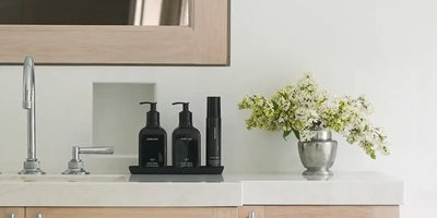 Luxury fragrance home products set displayed on a black tray beside white flowers on a marble bathroom counter.