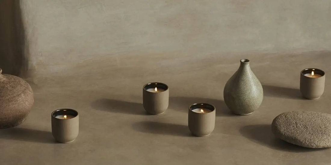 Luxury scented candles collection of four lit mini candles in ceramic vessels styled with stone and ceramic vases on a neutral surface.