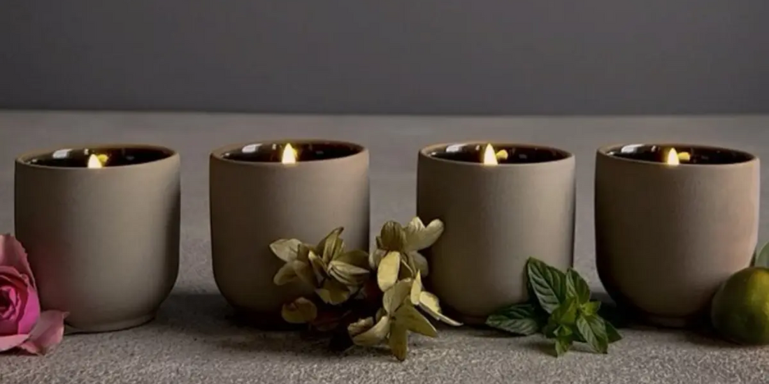 Four lit all-natural room deodorizer candles in minimalist holders, accompanied by a rose, dried flowers, mint leaves, and a small green fruit.