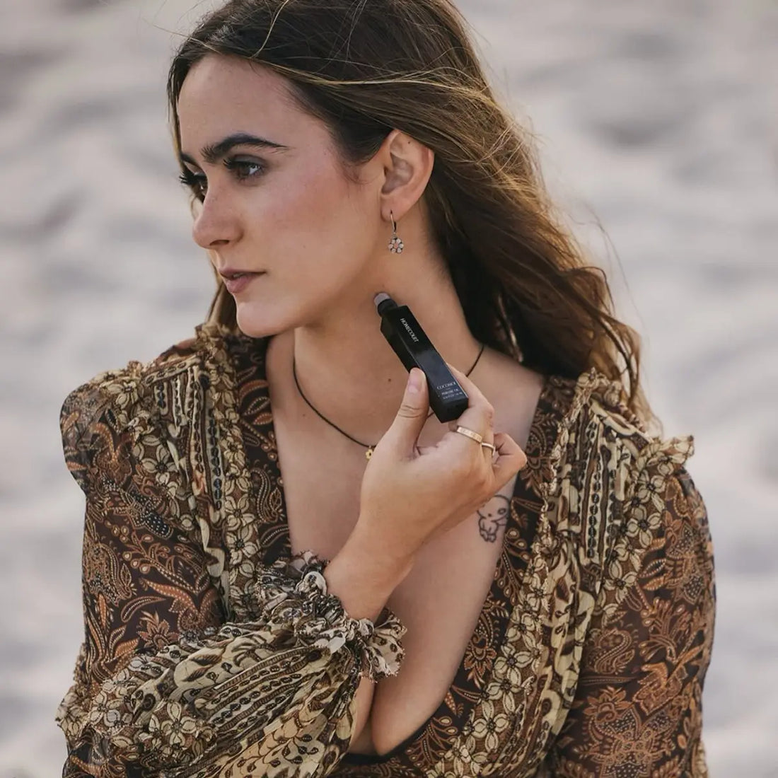 Woman applying non toxic perfume oil to her neck while sitting on a sandy beach, wearing a patterned dress.