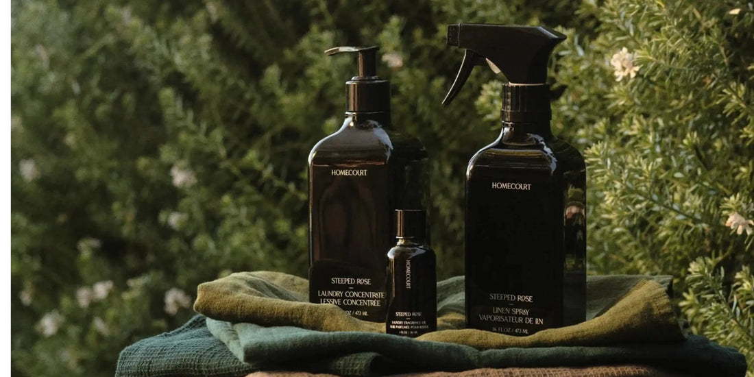 Homecourt Steeped Rose laundry concentrate, linen spray, and fragrance oil are arranged on folded textiles outdoors with lush greenery behind.