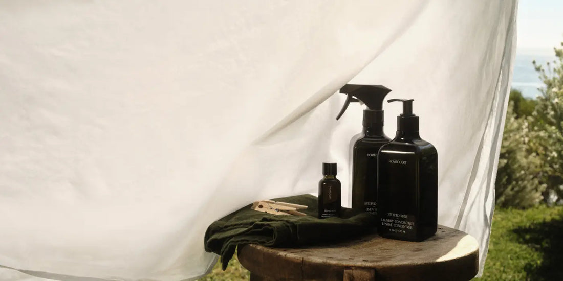 Non-toxic cleaners featuring elegant black bottles of eco-friendly laundry and linen sprays placed on a rustic stool outdoors.