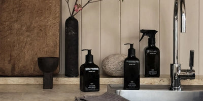 Homecourt organic all-purpose cleaner and hand wash bottles styled on a marble countertop beside a stone vase and chrome faucet.