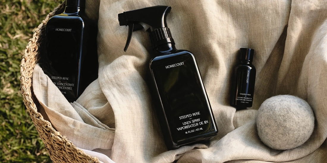 Homecourt Steeped Rose linen spray sits in a woven basket on beige linen with wool dryer balls on grass.