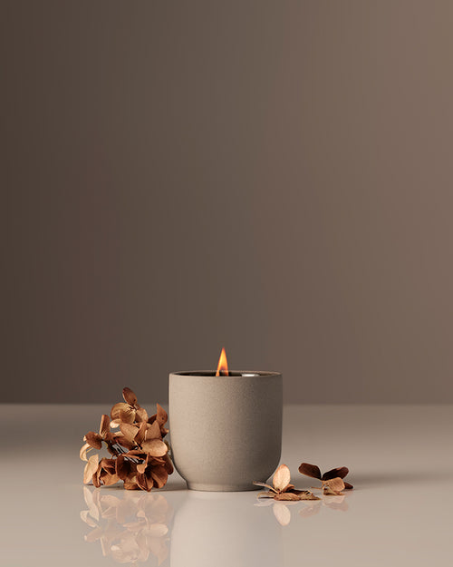 Candles in a beige vessel with dried floral accents on a reflective surface.