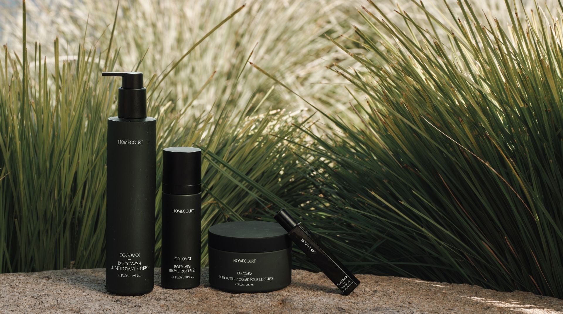 Four black skincare products from homecourt are arranged on a stone surface in front of tall green ornamental grass. The products include a pump bottle, a small bottle, a jar, and a tube, with white text on the labels.