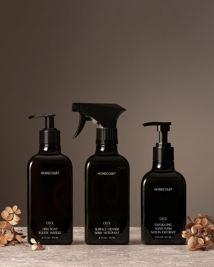 Homecourt Exfoliating Kitchen Trio in Cece fragrance, featuring Dish Soap, Surface Cleaner, and Exfoliating Hand Wash in black bottles arranged on a stone surface with dried florals against a warm neutral backdrop.